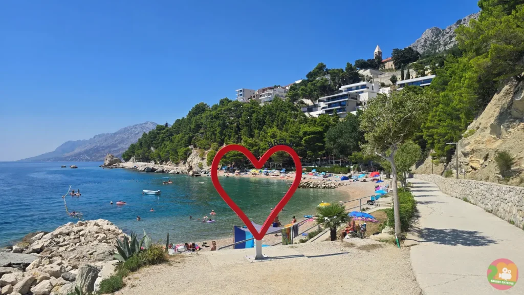 Crvena skulptura srca na šetnici u Breli iznad tirkizne uvale, iza nje borova šuma, hoteli i crkveni zvonik