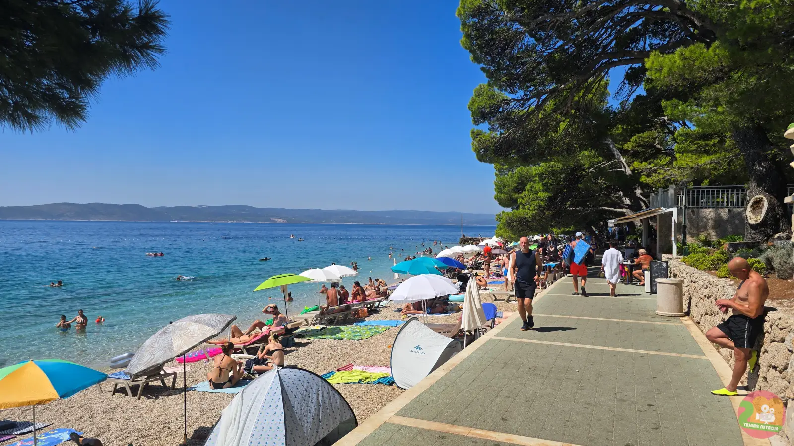 Puna šljunčana plaža uz šetnicu u Breli sa suncobranima, šatorima za plažu i kupačima u bistrom moru