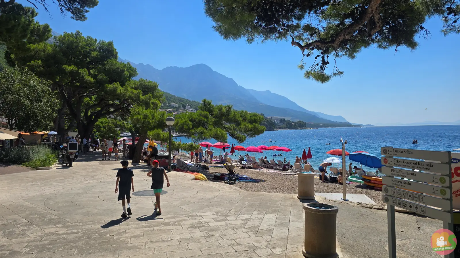 Popločani trg uz plažu Soline u centru Brele, s crvenim suncobranima, putokazom i Biokovom u pozadini
