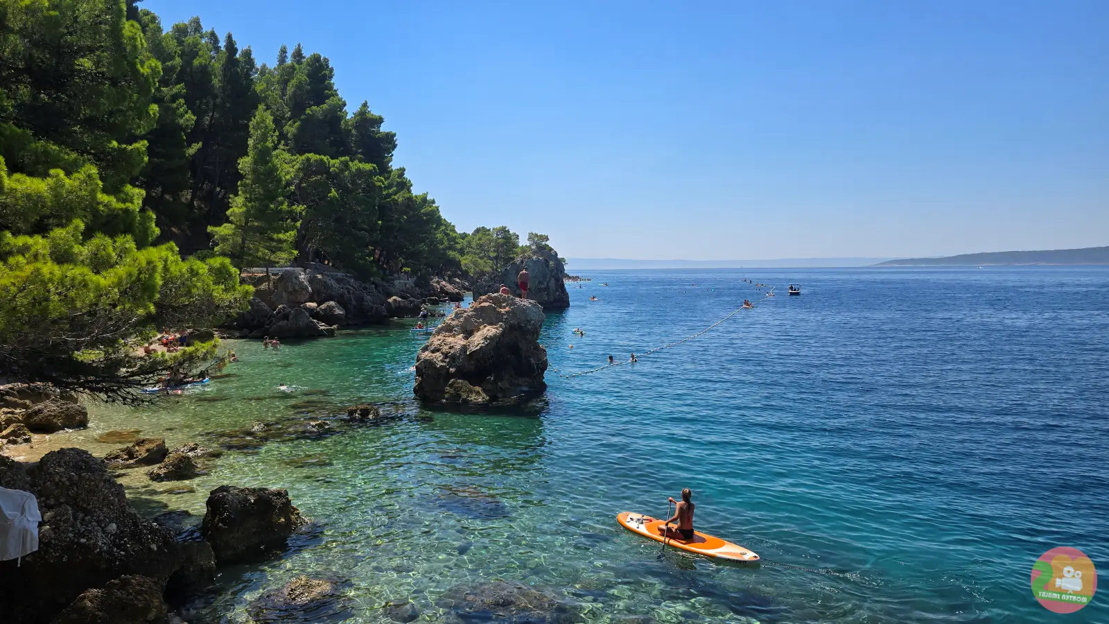 Felsige Küste mit Pinien in Brela, ein Stand Up Paddler auf orangem Board im klaren, türkisfarbenen Wasser